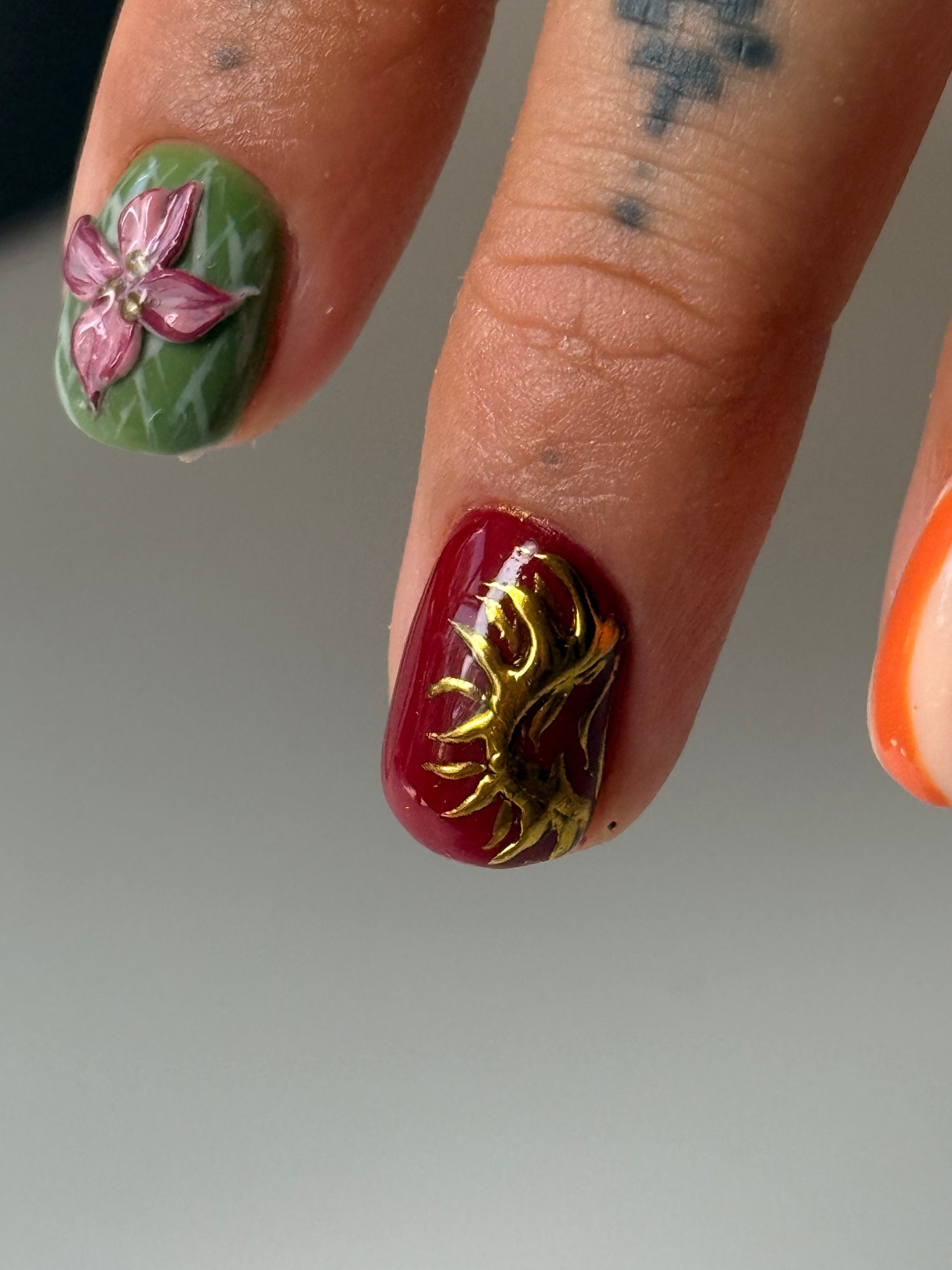 Close-up of a hand showing detailed nail art designs on three fingernails, including a green nail with a pink flower, a deep red nail with a gold sun motif, and a partial orange nail.